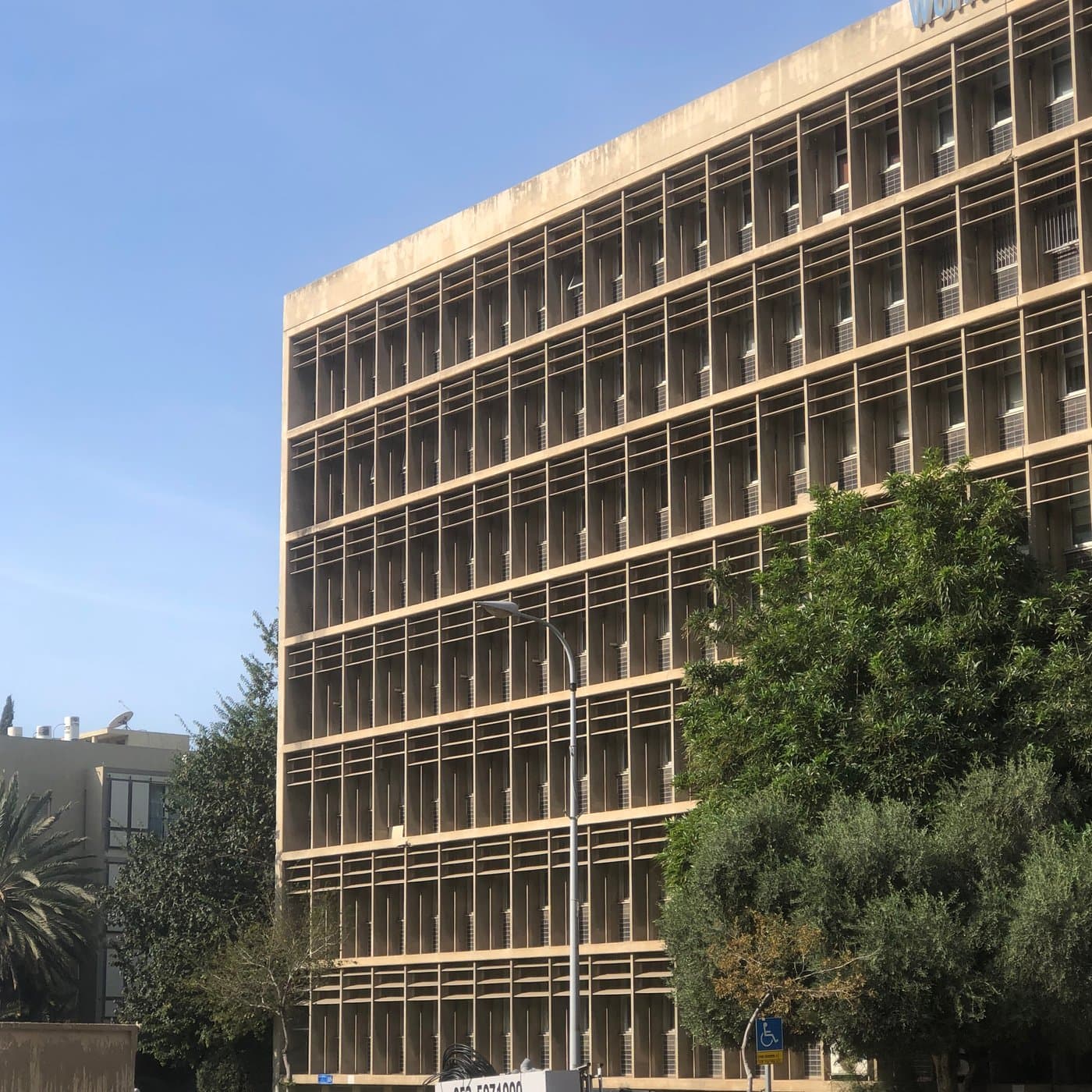 חזית ארוכה של בניין ברוטליסטי עם רשת חלונות ומצללות בטון חוזרות, עצים ופנס רחוב לפניו