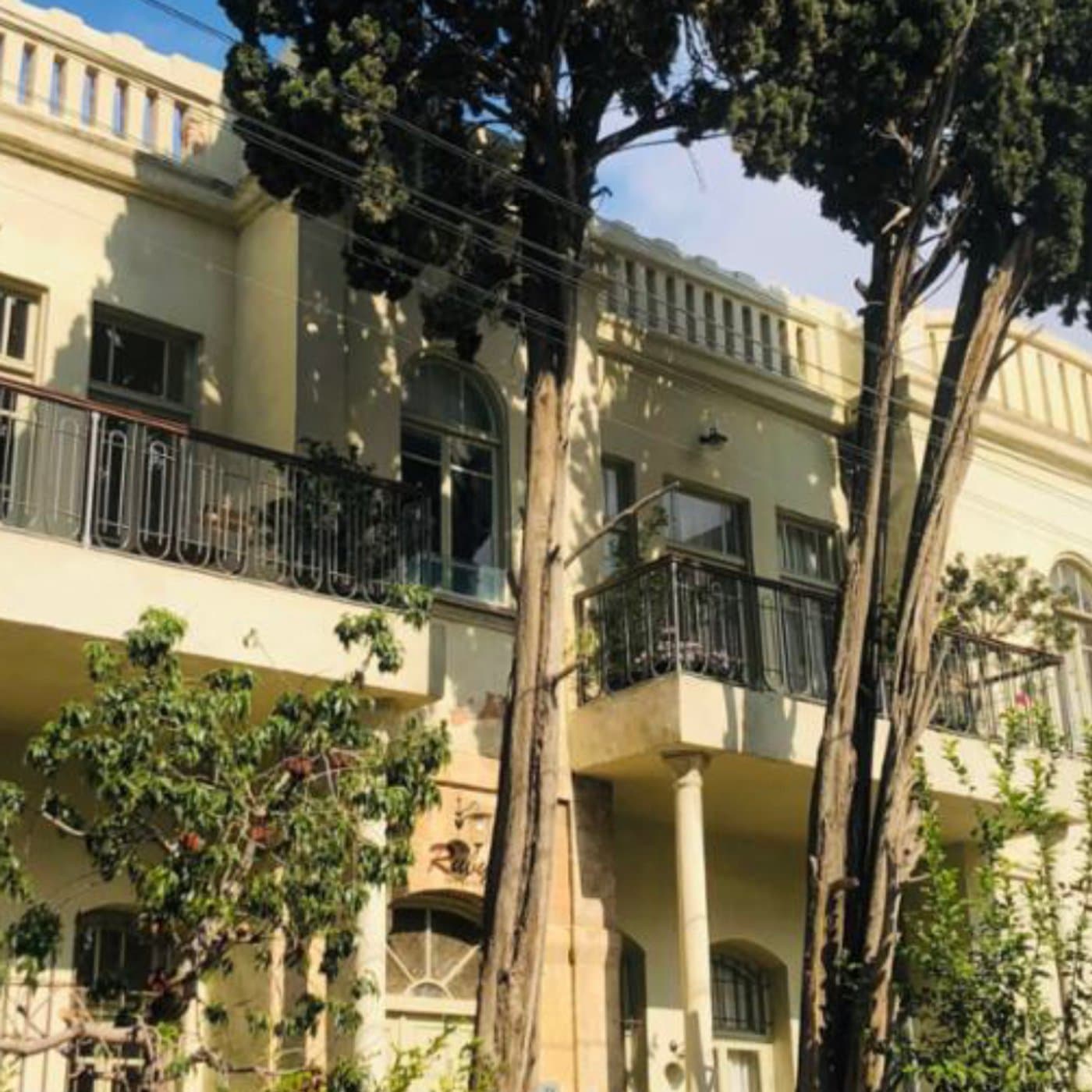 בניין אקלקטי בגוון קרם עם מעקה גג מסולסל וחלונות קשתיים, ברושים גבוהים לפניו