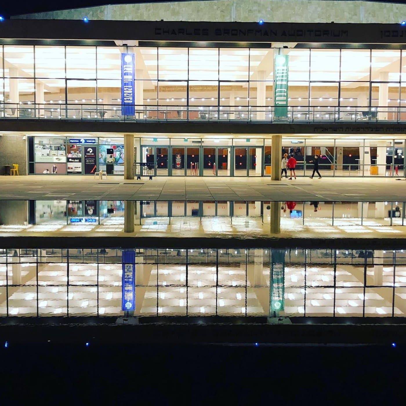 אודיטוריום צ'רלס ברונפמן מואר בלילה, חזית זכוכית עם דגלים תלויים, בריכת מים משתקפת מתחתיו