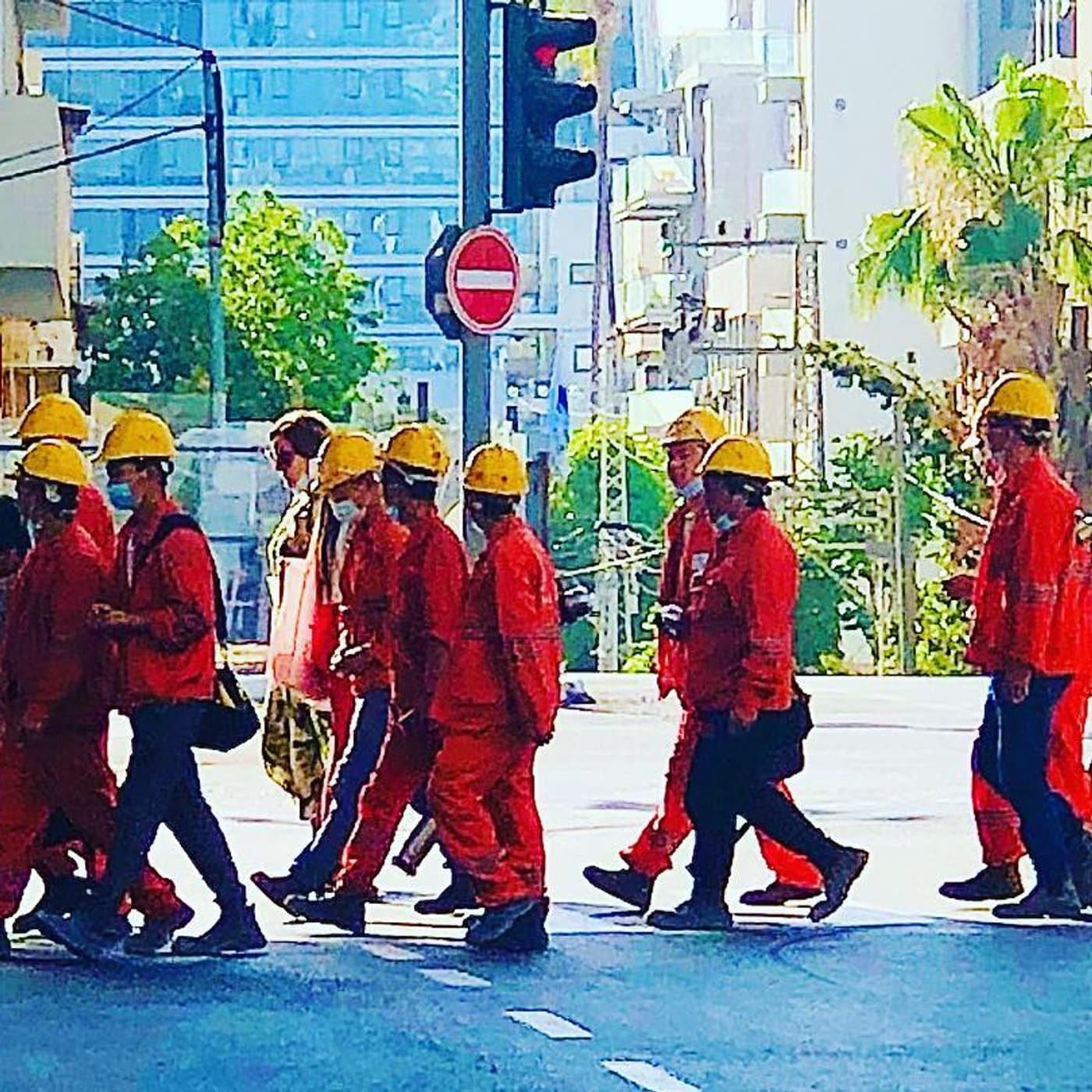 קבוצת פועלי בניין בסרבלים כתומים וקסדות צהובות חוצה כביש עירוני, רוכב אופניים ודקל ברקע
