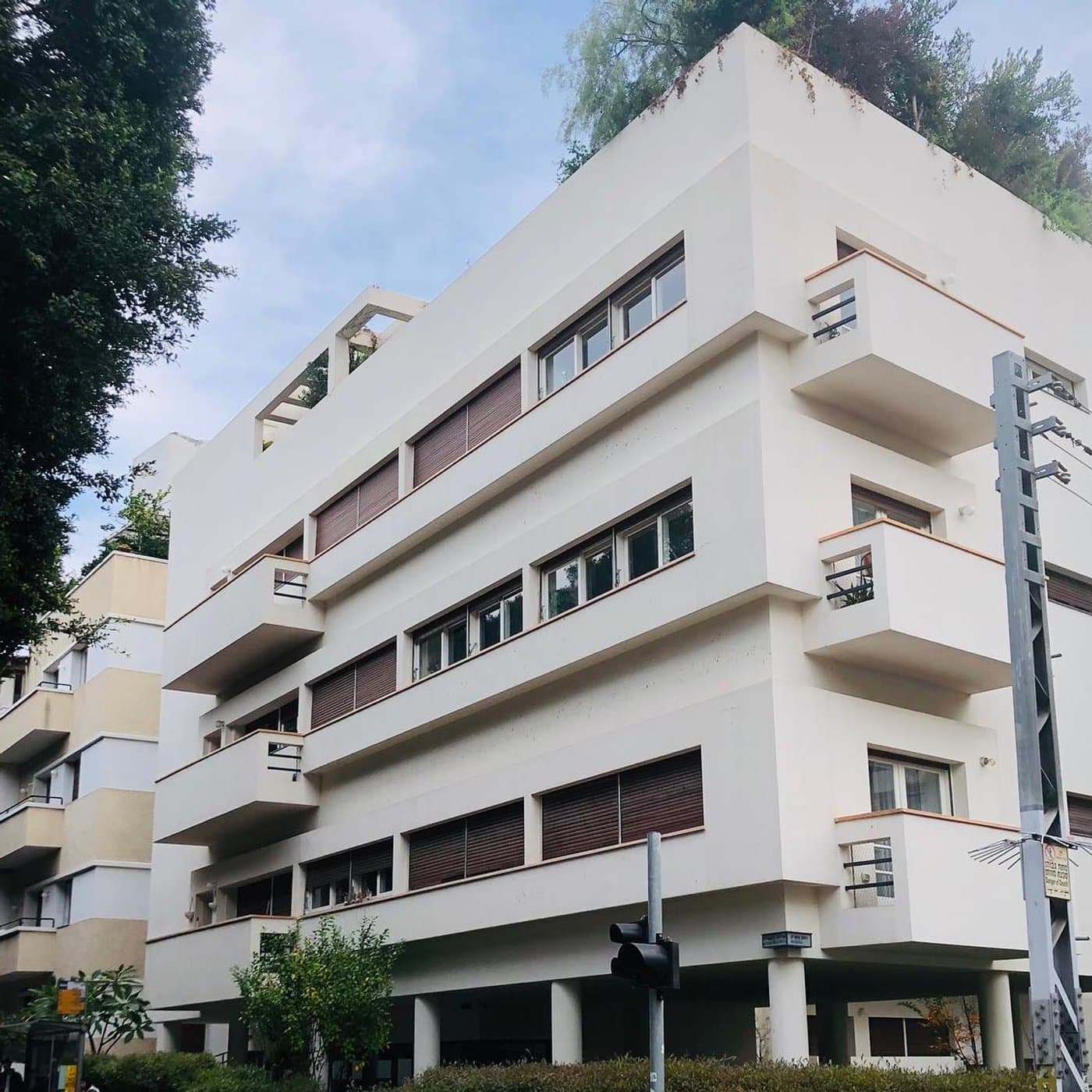 בניין באוהאוס קלאסי לבן העומד על עמודים, גינת גג ירוקה על הגג ופינת רחוב עם רמזורים