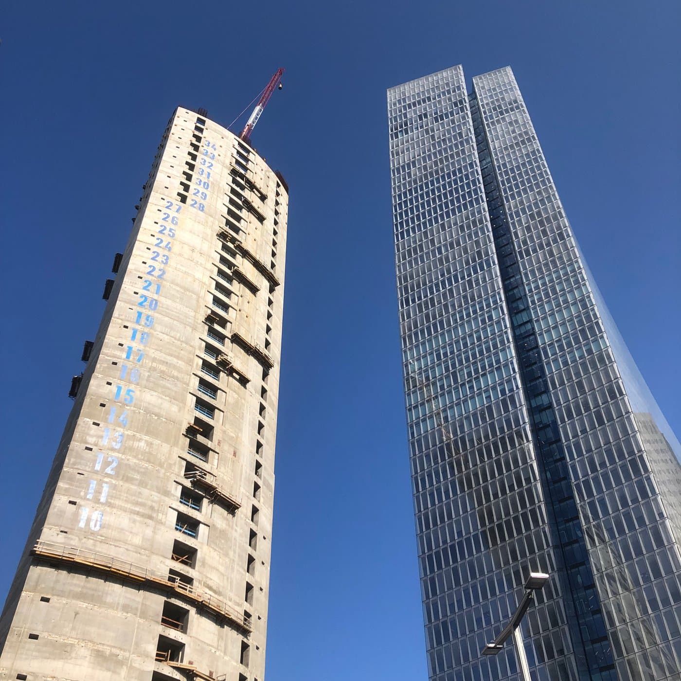 מגדל בטון גולמי בבנייה עם מספרי קומות צבועים על החזית ועגורן אדום, מגדל זכוכית גמור חוצה באלכסון מעליו