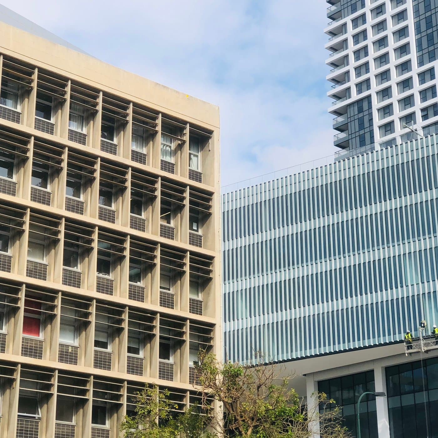 חזית ברוטליסטית עם רשת חלונות שקועים בבטון בגוון חול, מול מגדל זכוכית עכשווי ופועלים על במת עבודה תלויה