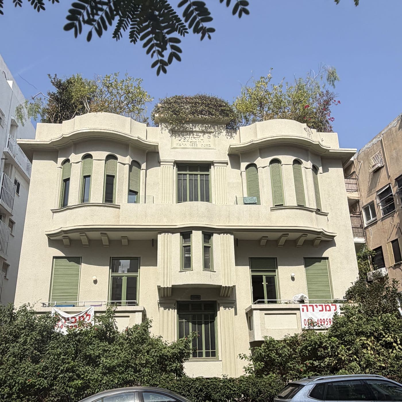בניין אקלקטי בהיר ברחוב פינסקר, חזית סימטרית עם תריסים ירוקים וצמחייה על הגג